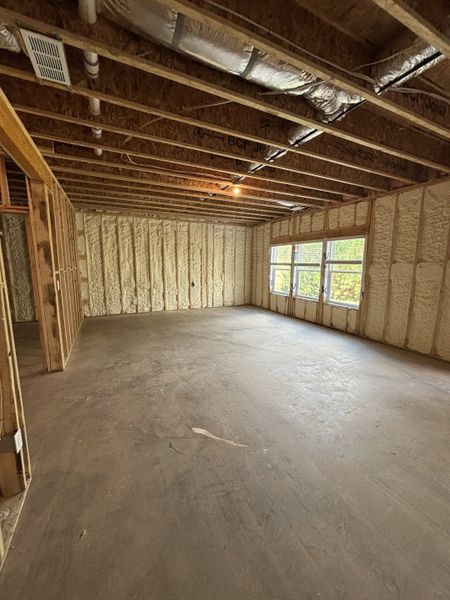 An unfinished room with exposed beams, spray foam insulation, and large windows for natural light.