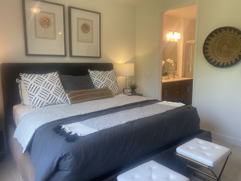 A cozy bedroom with a plush bed, decorative pillows, modern wall art, and an ensuite bath featuring elegant lighting and dark wood vanity.