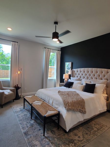 A cozy bedroom featuring a tufted headboard, modern ceiling fan, and natural light through large windows.