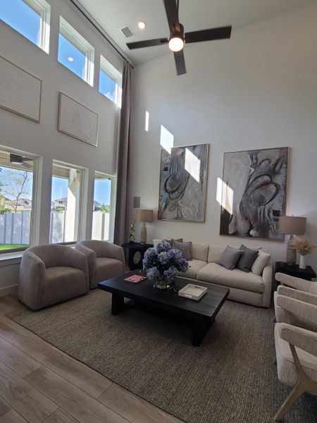 A cozy living room with high ceilings, large windows, abstract art, neutral furniture, and a modern ceiling fan.