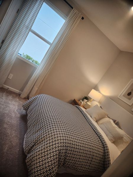 A cozy bedroom with a geometric-patterned bedspread, neutral walls, a soft carpet, and soft natural light filtering through curtains.