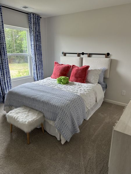 A cozy bedroom with plush red pillows, blue curtains, and a fluffy stool beneath a bright window.