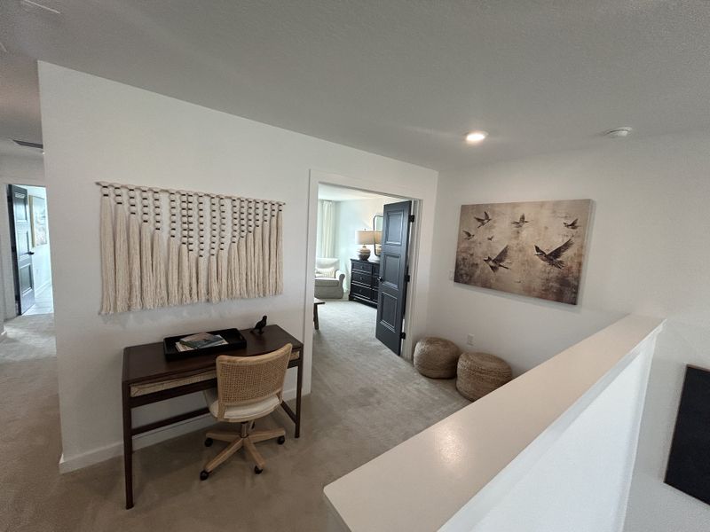 A cozy nook with a woven wall hanging, compact desk, and abstract bird artwork, leading to a bright room with plush seating.
