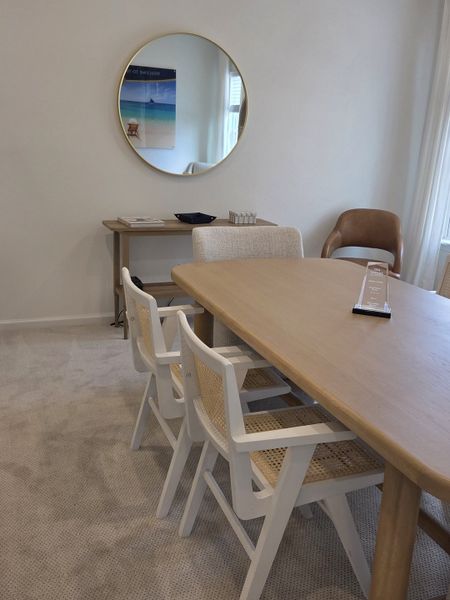 A cozy dining area featuring a wooden table, white woven chairs, and a round mirror on a neutral carpet.