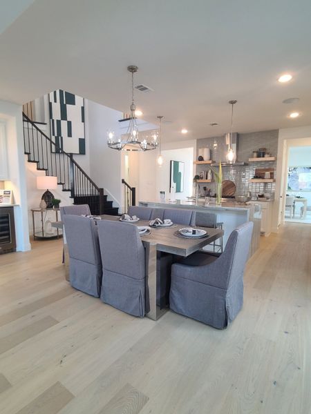 A modern dining area with a chic chandelier, grey upholstered chairs, and open shelving, leading to a stylish kitchen.