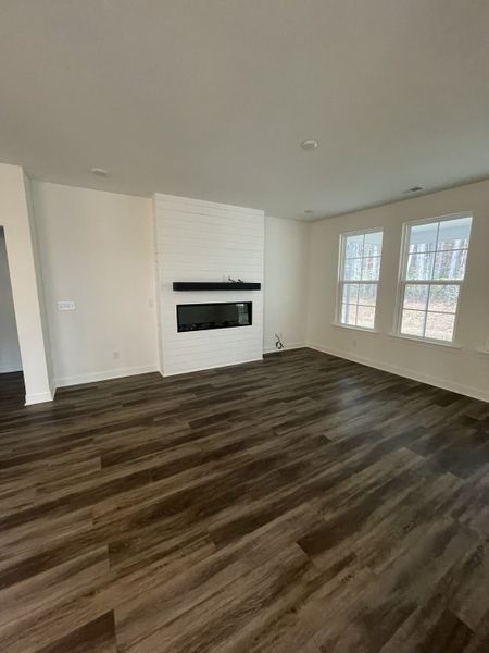 A spacious living area with warm wood flooring, stylish white shiplap fireplace, and abundant natural light through large windows.