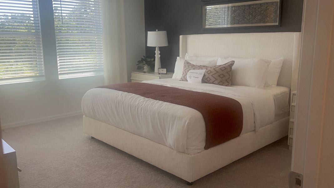 A cozy bedroom with a plush white bed, accented by a rust throw, soft lighting, and natural light from large windows.