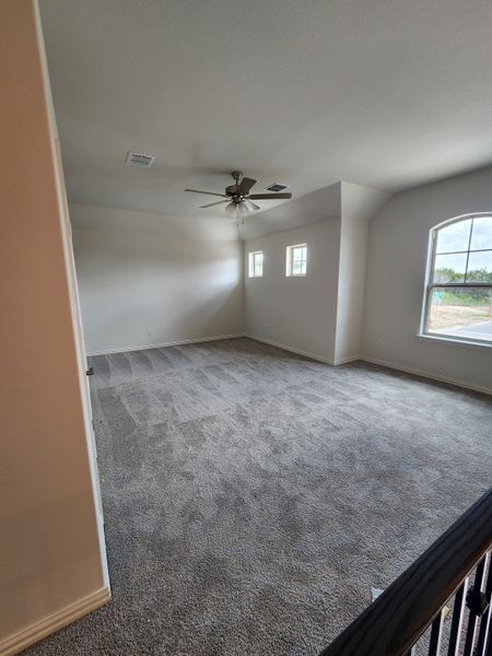 A spacious carpeted room with neutral walls, large windows, and a ceiling fan, offering ample natural light and comfort.