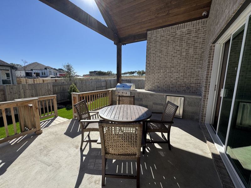 A cozy patio with a round table, woven chairs, and a built-in grill under a wooden pergola. Ideal for outdoor relaxation.