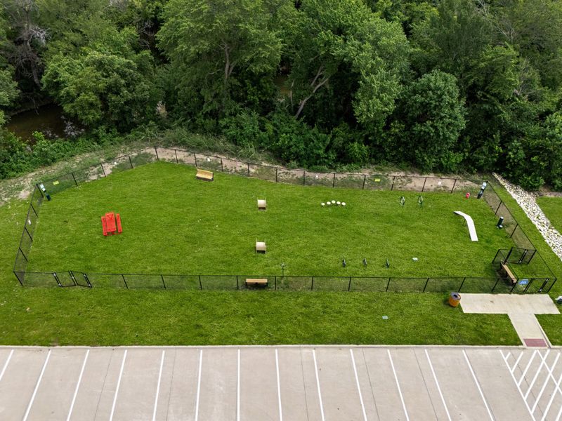 Aerial view of fenced dog park with agility equipment on green grass, next to a parking lot and wooded area Aerial view of fenced dog park with agility equipment on green grass, next to a parking lot and wooded area