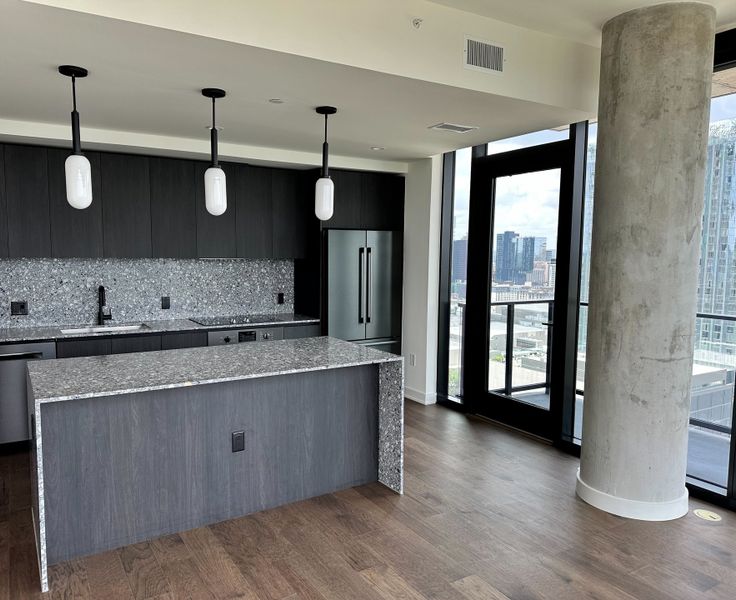A modern kitchen with sleek dark cabinetry, terrazzo countertops, pendant lighting, and expansive city views through large windows.