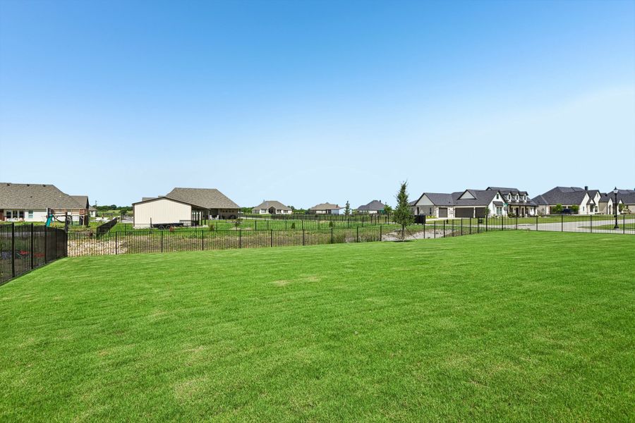 Large backyard with green lawn, black iron fence, and neighboring homes visible in the distance. Large backyard with green lawn, black iron fence, and neighboring homes visible in the distance.