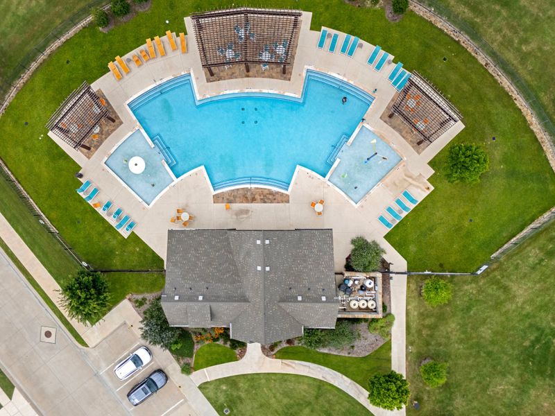 Aerial view of community pool complex with lounge chairs, pergola, and clubhouse building surrounded by green lawn Aerial view of community pool complex with lounge chairs, pergola, and clubhouse building surrounded by green lawn