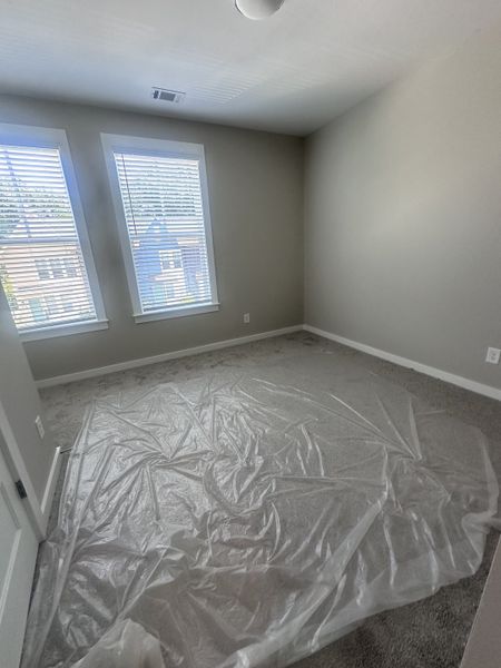 A bright bedroom with natural light from two windows, neutral walls, and carpeted flooring covered in protective plastic.