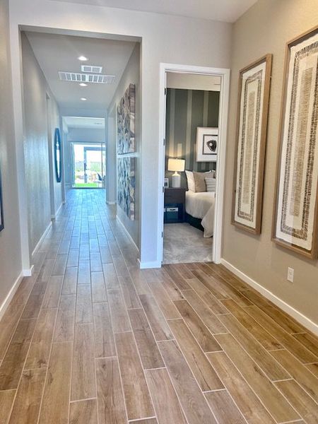 A modern hallway with wood-like tile flooring, elegant artwork, and a view into a cozy, well-lit bedroom.