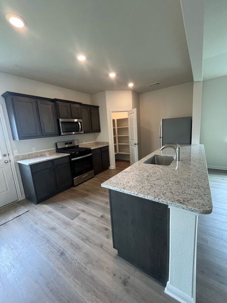 A modern kitchen with granite countertops, dark cabinets, wood flooring, and recessed lighting, featuring a spacious pantry.