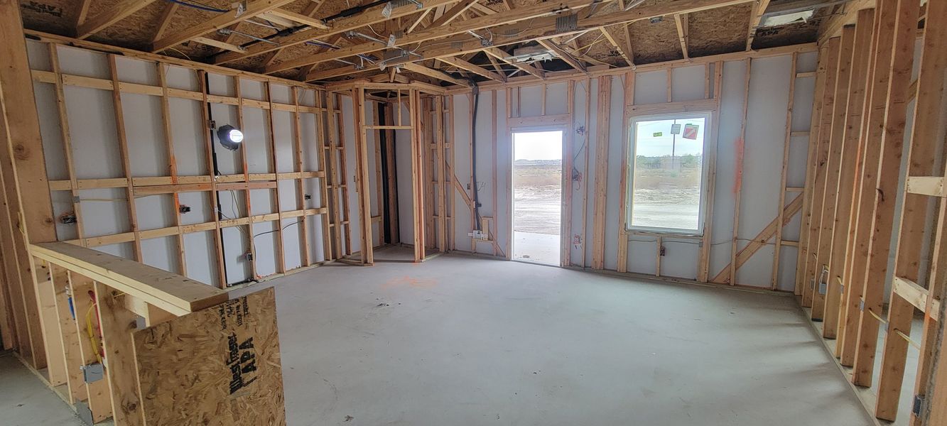 A construction interior with exposed wooden framing and wiring, featuring a concrete floor and natural light from large windows.