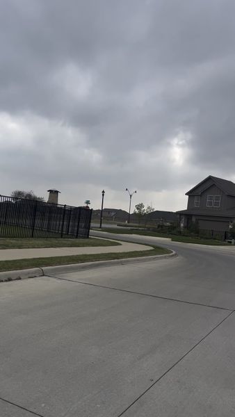 A serene street view in Enchanted Creek by Our Country Homes, Lucas, TX, featuring a modern home under cloudy skies.