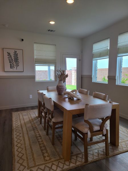 A cozy dining room with a wooden table, cushioned chairs, and natural light from wide windows, creating a warm ambiance.