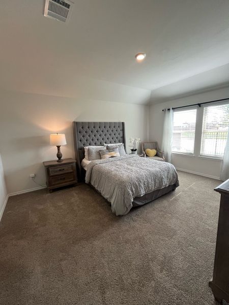 A cozy bedroom with a tufted headboard, elegant bedding, soft carpeting, and natural light filtering through large windows.