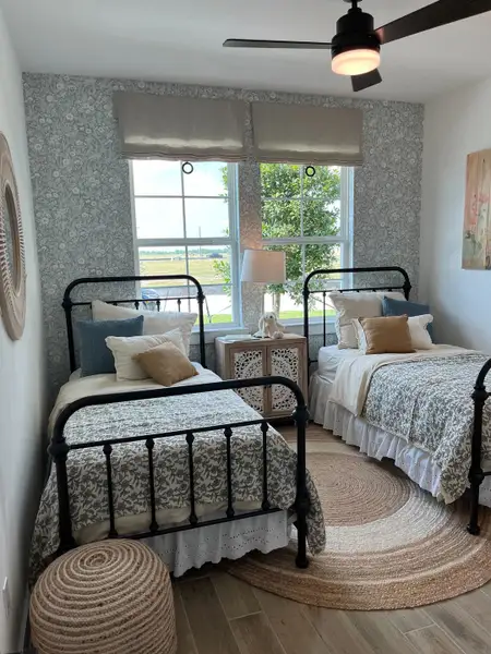 A cozy bedroom featuring twin metal beds, floral bedding, a woven rug, and soft natural light from large windows.