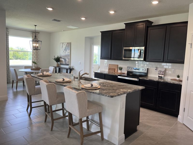 A modern kitchen with dark cabinets, a granite island, beige seating, and elegant lighting, adjacent to a bright dining area. A modern kitchen with dark cabinets, a granite island, beige seating, and elegant lighting, adjacent to a bright dining area.