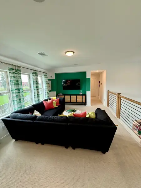 A cozy living room featuring a dark sectional sofa, vibrant cushions, and a TV, accented by green walls and airy windows.