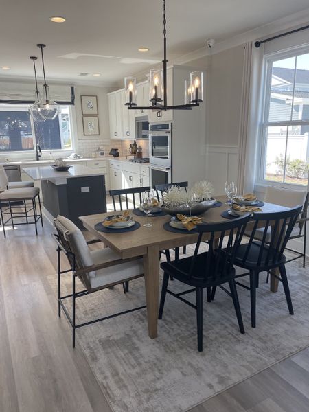 A stylish dining area with a wooden table set for six, modern black chairs, a bright kitchen with white cabinets, and large windows bringing in natural light.