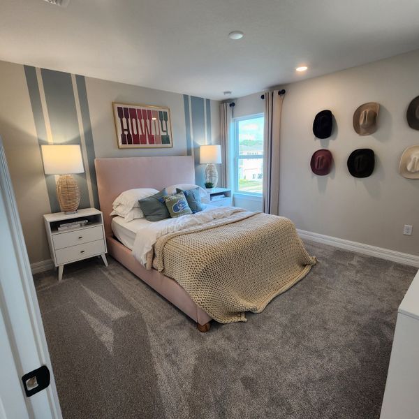 A cozy bedroom with a plush bed, accent wall, and hat decor. Warm lighting and carpeted flooring create a welcoming ambiance.