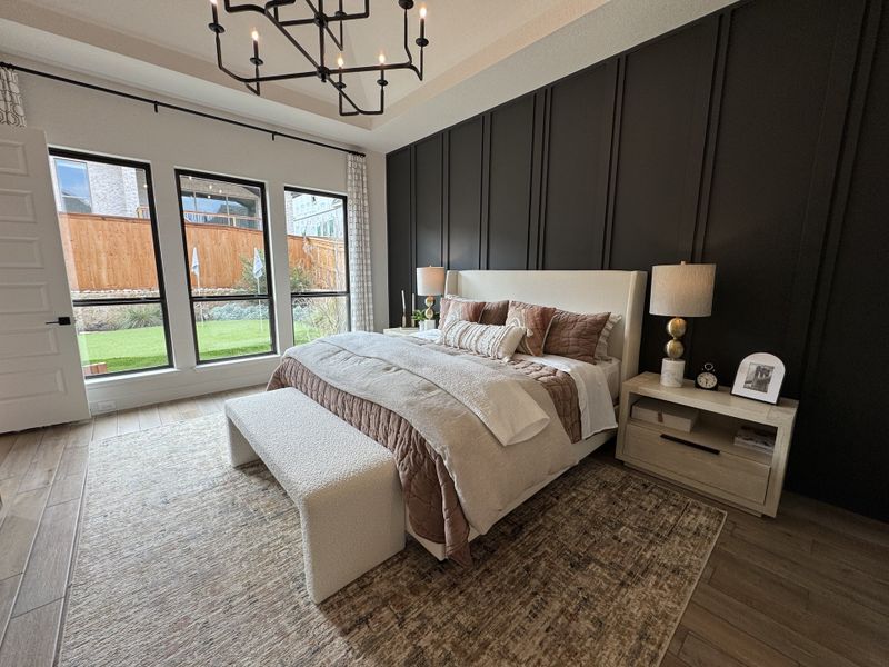 A cozy bedroom with a dark accent wall, large windows, chic lighting, and a textured bench at the foot of the plush bed.