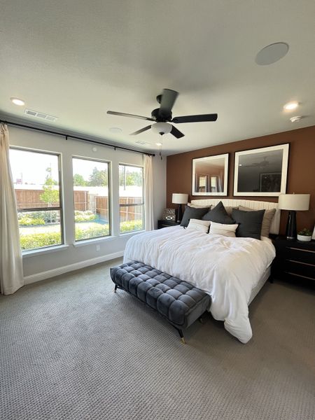 A cozy bedroom featuring large windows, a stylish ceiling fan, and a plush bench at the foot of the bed.