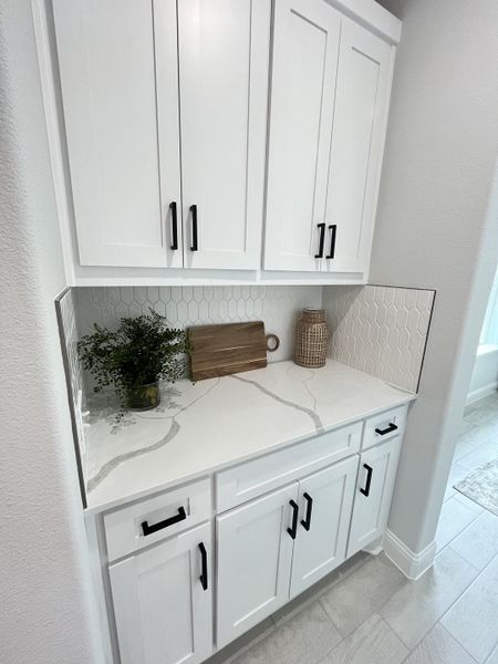 Sleek kitchen nook with white cabinetry, hexagonal backsplash, and black hardware, featuring stylish decor elements.