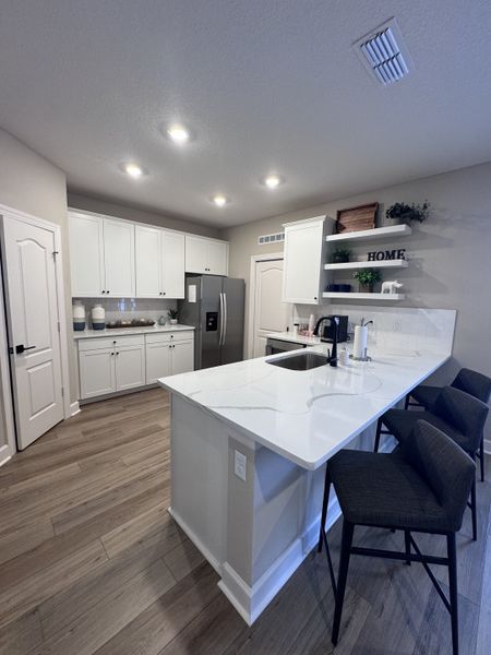 A sleek kitchen with white cabinets, quartz countertops, and dark bar stools, featuring modern appliances and wood flooring.