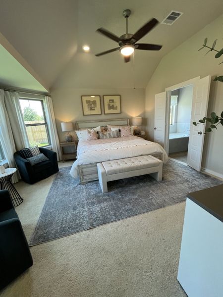 A cozy bedroom with a vaulted ceiling, ceiling fan, and elegant decor featuring a plush bed, armchairs, and large windows.