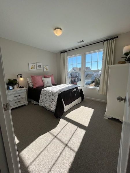 A cozy bedroom with a large window, soft carpeting, and bright decor, creating a welcoming and airy atmosphere.