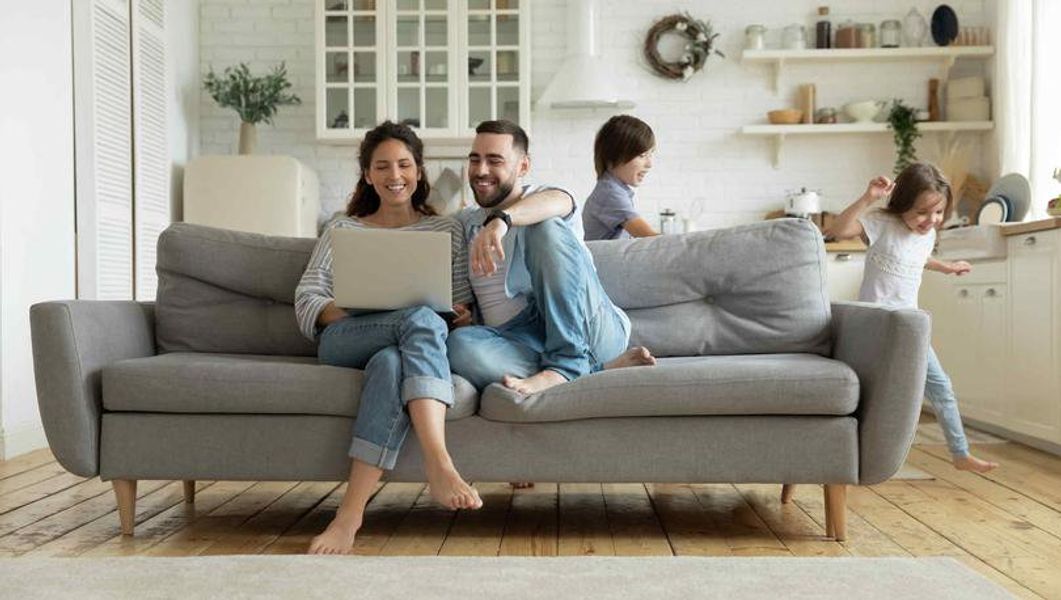 Couple sitting on a gray couch looking at a laptop, while two children play in the room.