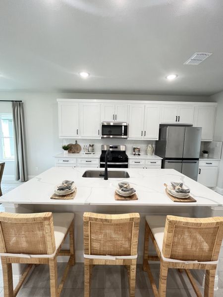 A modern kitchen with white cabinetry, an elegant island, wicker chairs, and stainless steel appliances.