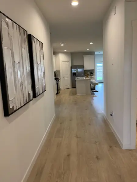 A modern hallway with light wood floors, abstract art, and view into a bright kitchen with contemporary design.