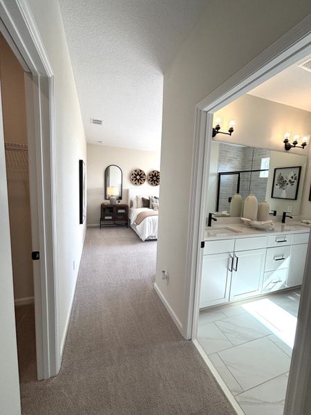 A bright hallway with carpeted floors leads to a bedroom and a bathroom featuring a sleek double vanity and modern fixtures.