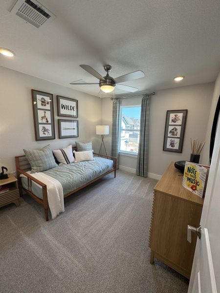 A cozy bedroom with a modern fan, daybed, stylish decor, and soft carpet, accented by framed art and natural light.