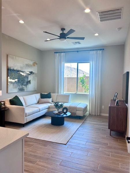 A cozy living room featuring a sectional sofa, modern ceiling fan, and large artwork, bathed in natural light.