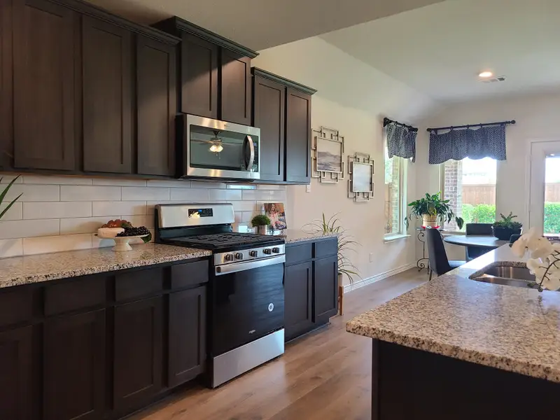 A modern kitchen with dark wood cabinets, granite countertops, and stainless steel appliances, leading to a bright dining area.
