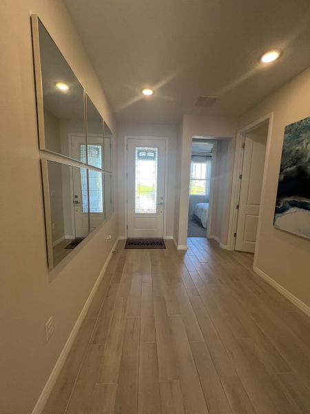 A bright entryway with light wood flooring, modern mirrors, and soft lighting, leading to a cozy bedroom space.