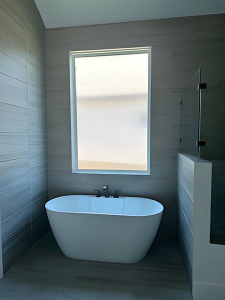 A serene bathroom with a sleek freestanding bathtub, floor-to-ceiling tiles, and ample natural light from a large frosted window.