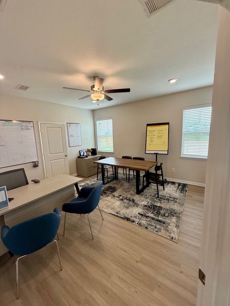A modern office space featuring sleek wooden desks, blue chairs, and a ceiling fan on a decorative rug.