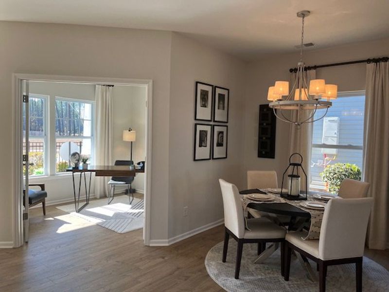 An elegant dining area with a round table and modern chandelier, adjacent to a bright home office with large windows.