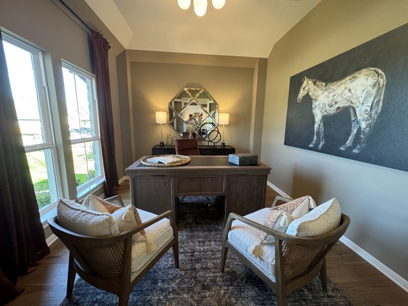 A cozy office with a wooden desk, plush chairs, and horse artwork, bathed in natural light from large windows.