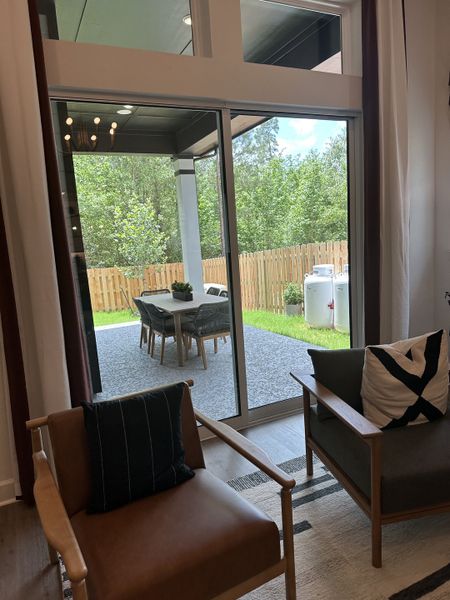 A cozy living area with modern chairs, large windows, and a view of a stylish patio set against a lush backyard.