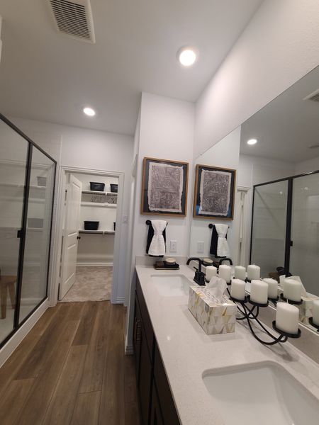 A modern bathroom featuring dual sinks, elegant wood flooring, a large glass shower, and spacious closet access.
