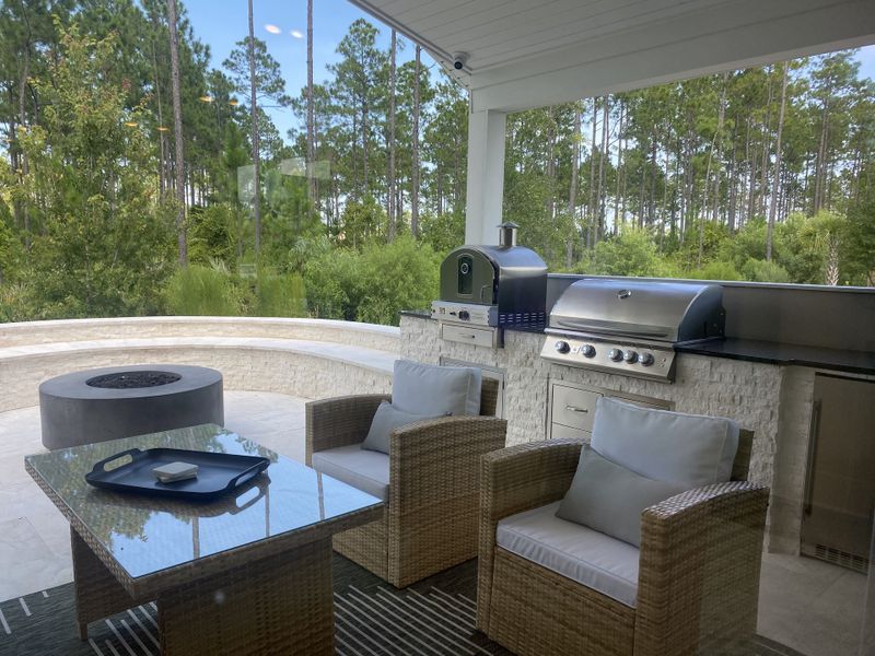 A modern outdoor patio with a built-in grill, pizza oven, wicker furniture, and serene woodland views.
